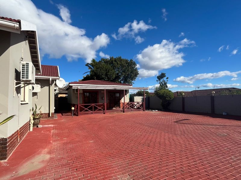A spacious rear exterior area featuring a large red paved driveway, a covered patio with wooden railings, and a secure perimeter wall.
