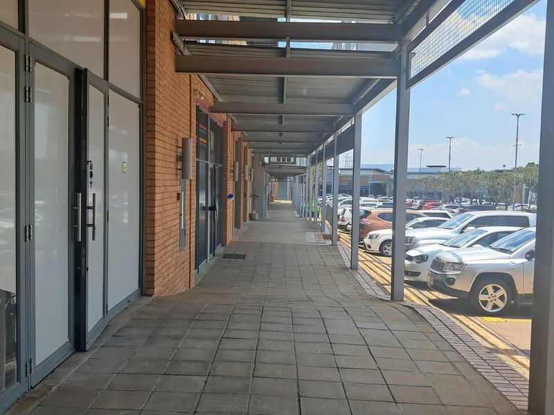 A covered exterior walkway featuring a steel canopy structure, paved flooring, and glass storefront entrances along a brick-faced industrial or commercial building.