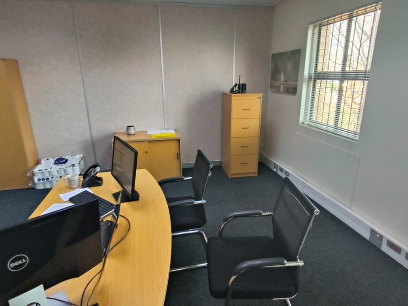 A functional office space featuring carpeted flooring, a window with blinds, and integrated HVAC unit, suitable for a professional work environment.