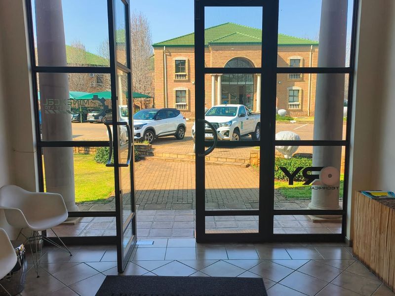 Bright office entrance featuring glass double doors, tiled flooring, and structural columns, providing a clear view of the exterior parking area.