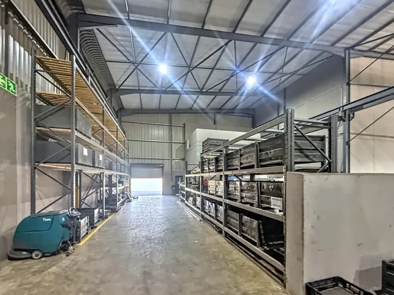 Spacious industrial warehouse area featuring high ceilings, steel portal frame construction, concrete flooring, and extensive fixed industrial shelving for storage.