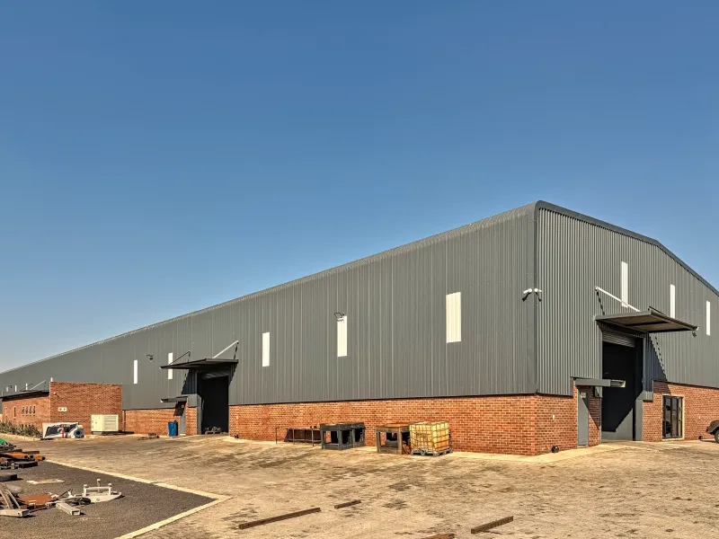 A large industrial warehouse facility featuring corrugated metal cladding, face brick lower walls, multiple roller shutter access points, and a spacious paved yard area.