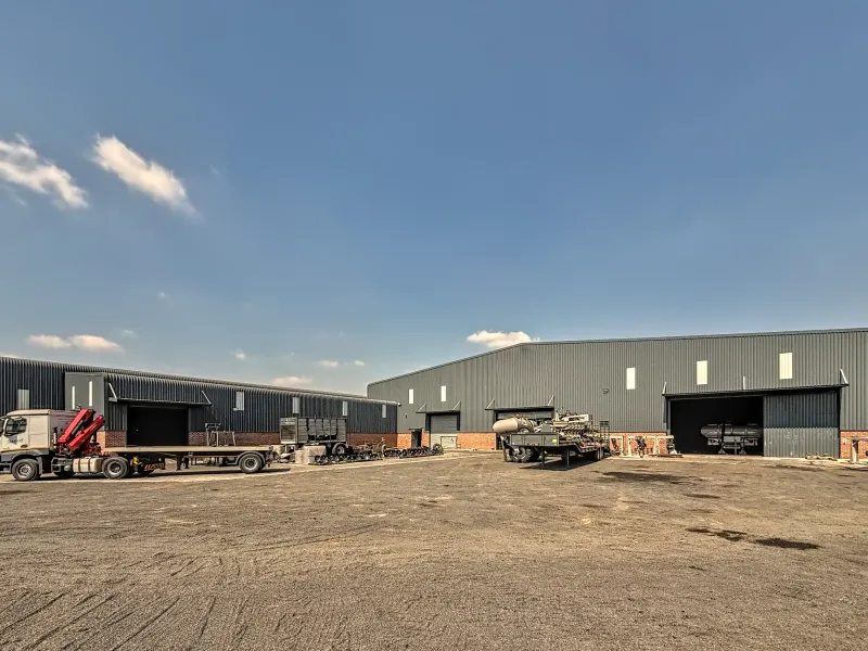 Spacious industrial yard featuring large metal-clad warehouse buildings with multiple roller shutter access points and a durable unpaved surface for heavy vehicle operations.