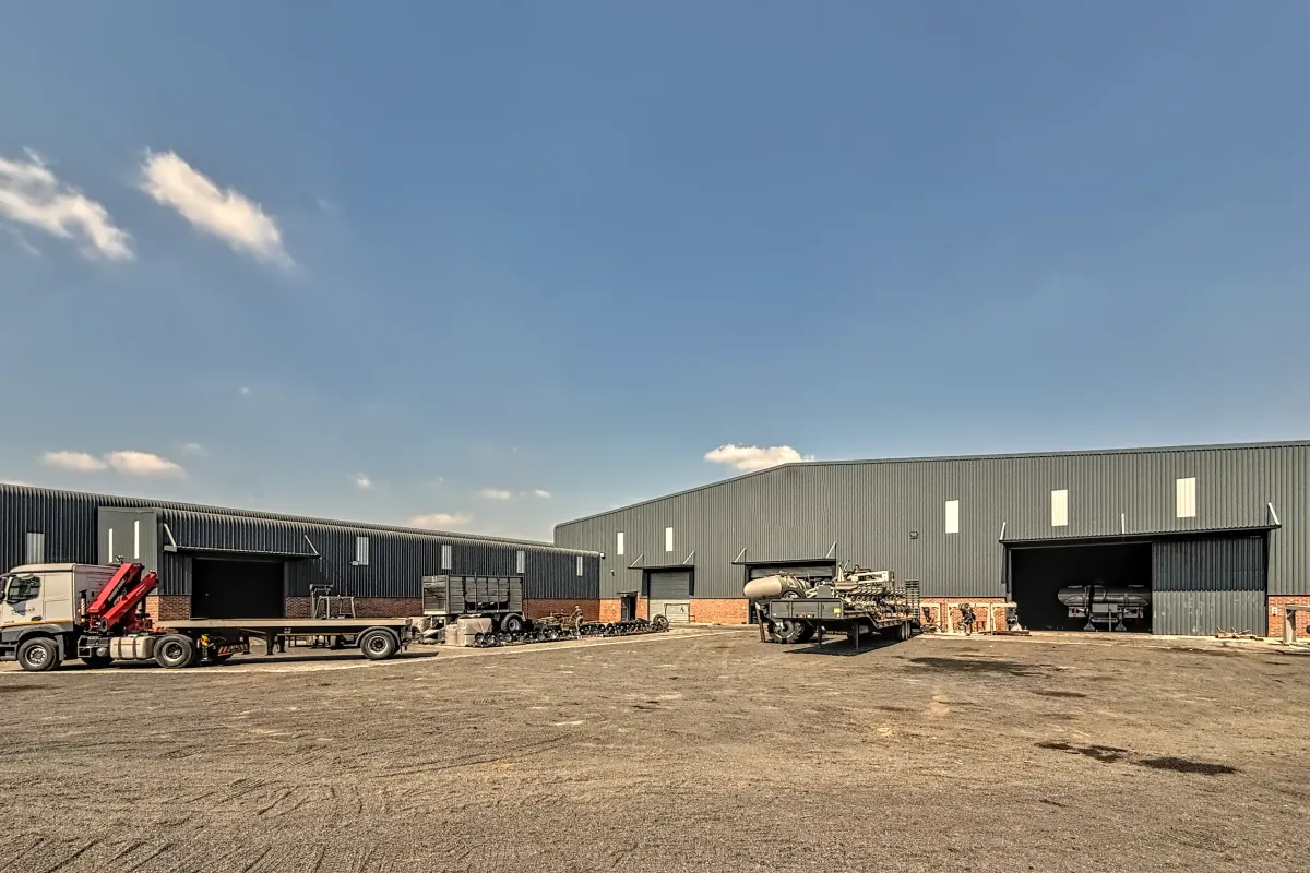 Spacious industrial yard featuring large metal-clad warehouse buildings with multiple roller shutter access points and a durable unpaved surface for heavy vehicle operations.