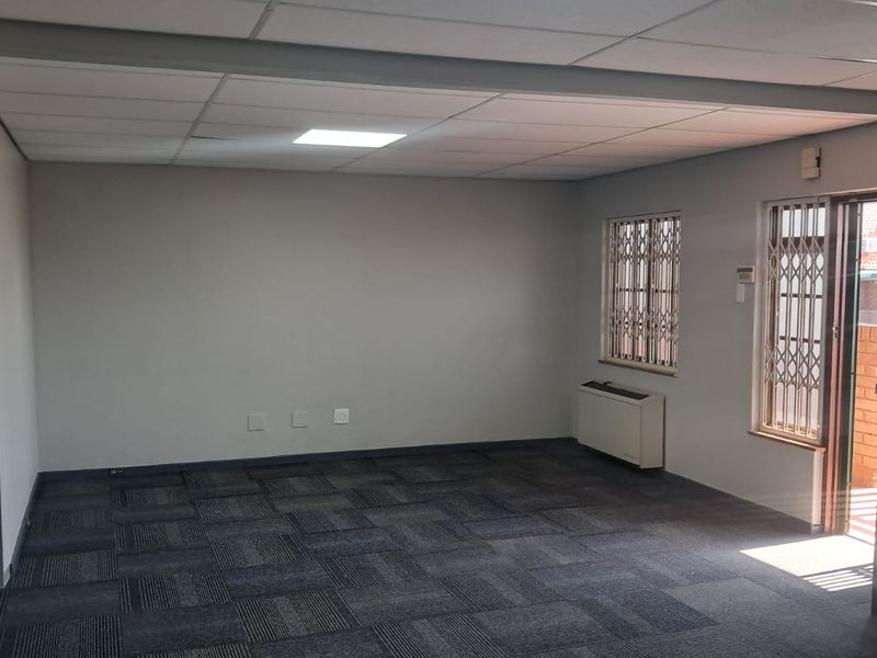A clean, empty office space featuring carpet tiles, a suspended ceiling with recessed lighting, air conditioning, and secure access via a door with security gates.