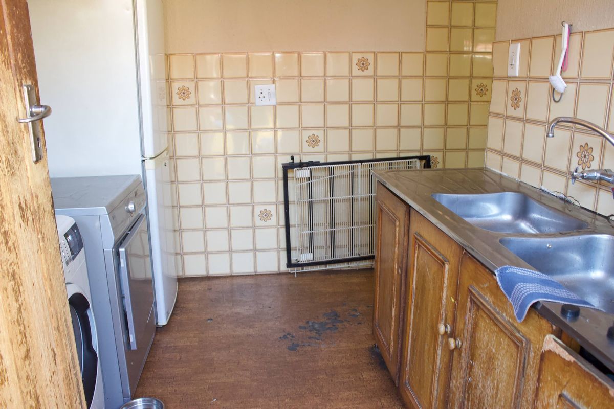 A compact kitchen area featuring dated wooden cabinetry, a double stainless steel sink, and decorative tiled splashbacks with worn flooring.