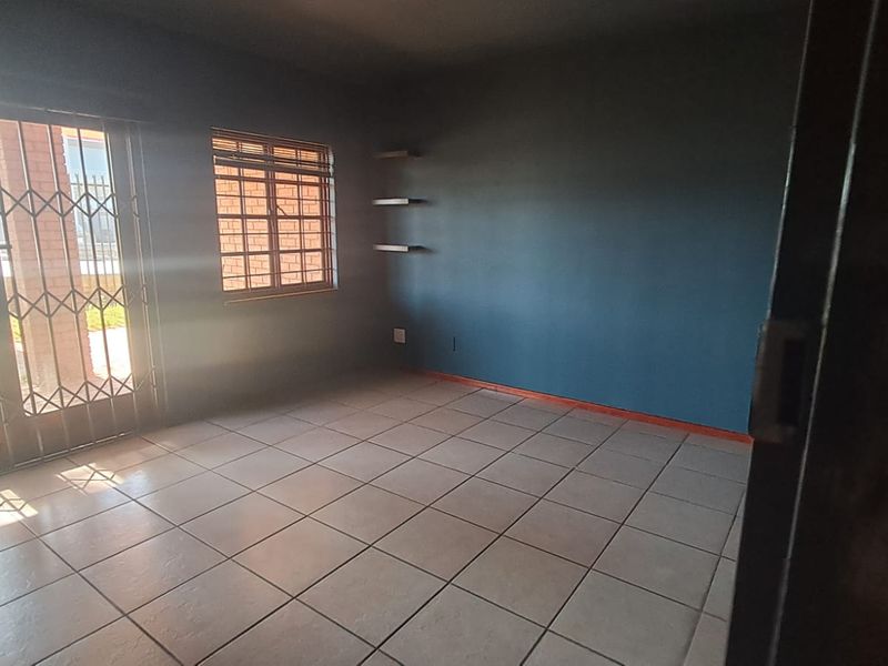 An empty office space featuring tiled flooring, a sliding door with security gate, and natural light from a window with burglar bars.