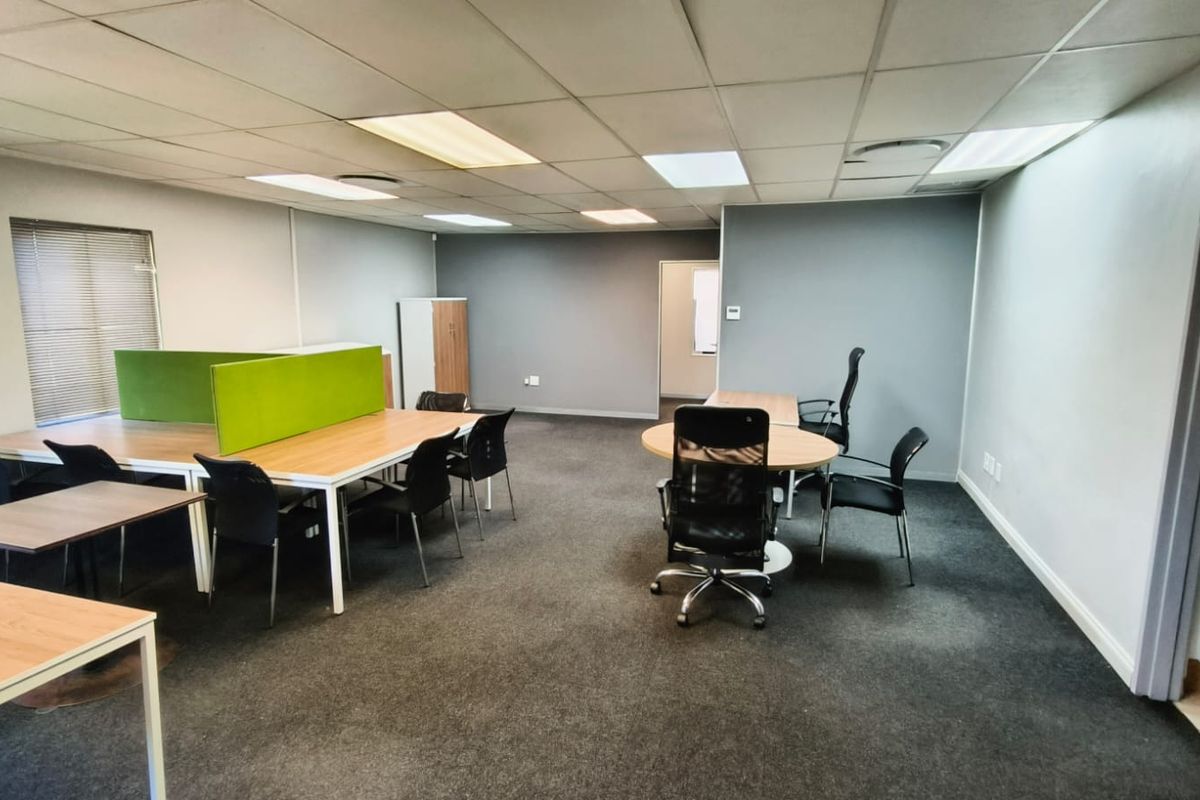 A functional office space featuring carpeted flooring, a suspended ceiling with recessed lighting, and large windows with venetian blinds.