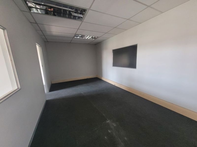 An empty office space featuring a suspended ceiling with recessed lighting, dark carpet flooring, and a wall-mounted whiteboard.