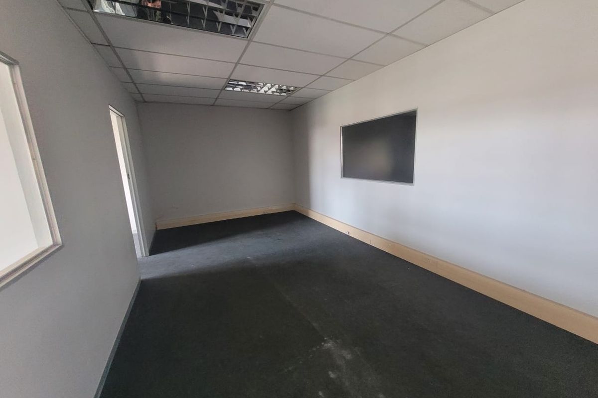 An empty office space featuring a suspended ceiling with recessed lighting, dark carpet flooring, and a wall-mounted whiteboard.