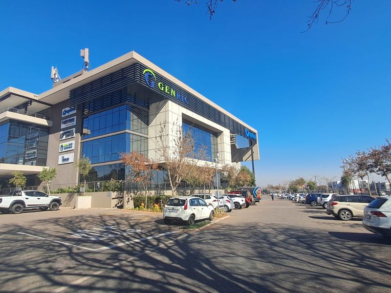 A modern multi-story commercial office building featuring a glass facade, prominent signage, and an extensive paved parking area.