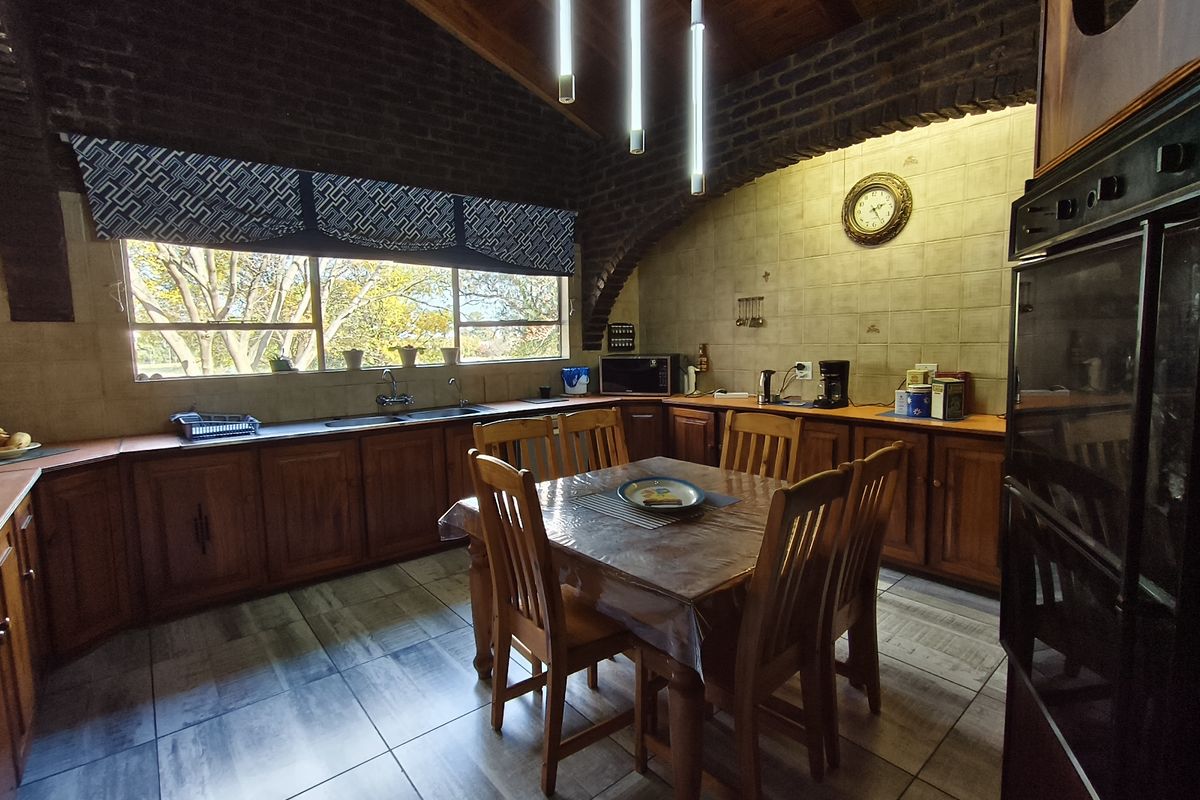 Spacious farmhouse kitchen featuring warm wooden cabinetry, exposed brick architectural details, timber ceiling beams, and a large window overlooking the garden.