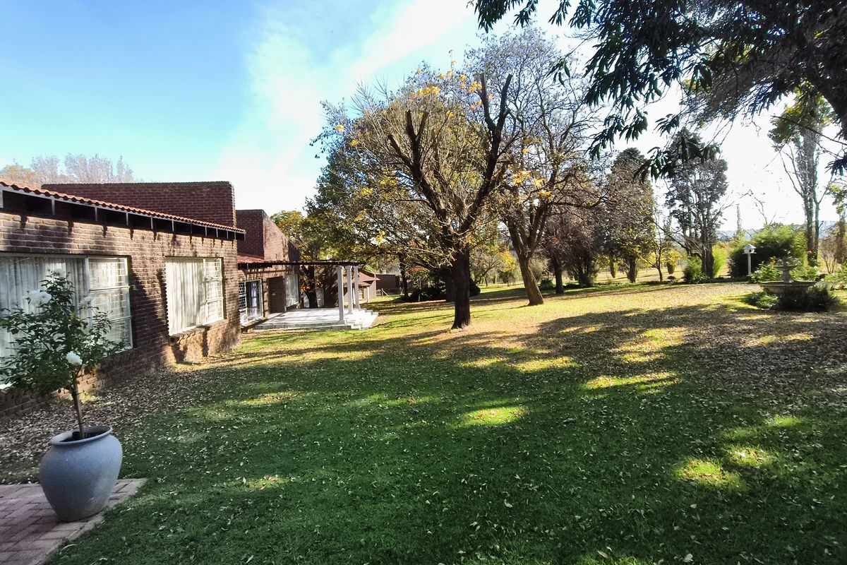 A view of the rear brick facade of the farmhouse, featuring a covered patio area and a spacious, well-maintained garden with mature trees.