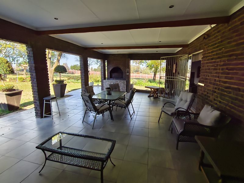 Spacious covered patio featuring a built-in braai, tiled flooring, and exposed brick pillars, offering a comfortable outdoor entertainment area overlooking the garden.