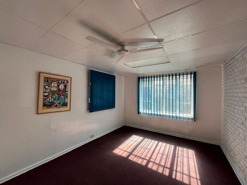 A vacant office space featuring a drop ceiling, ceiling fan, vertical blinds, and an exposed brick wall with carpet flooring.