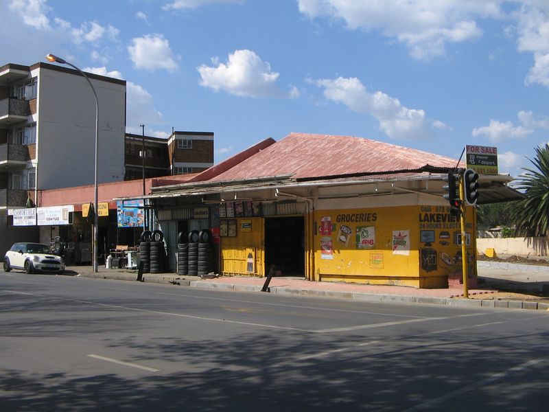 Street-level view of a retail property featuring a corrugated metal roof, yellow painted facade, and integrated security shutters along the sidewalk.