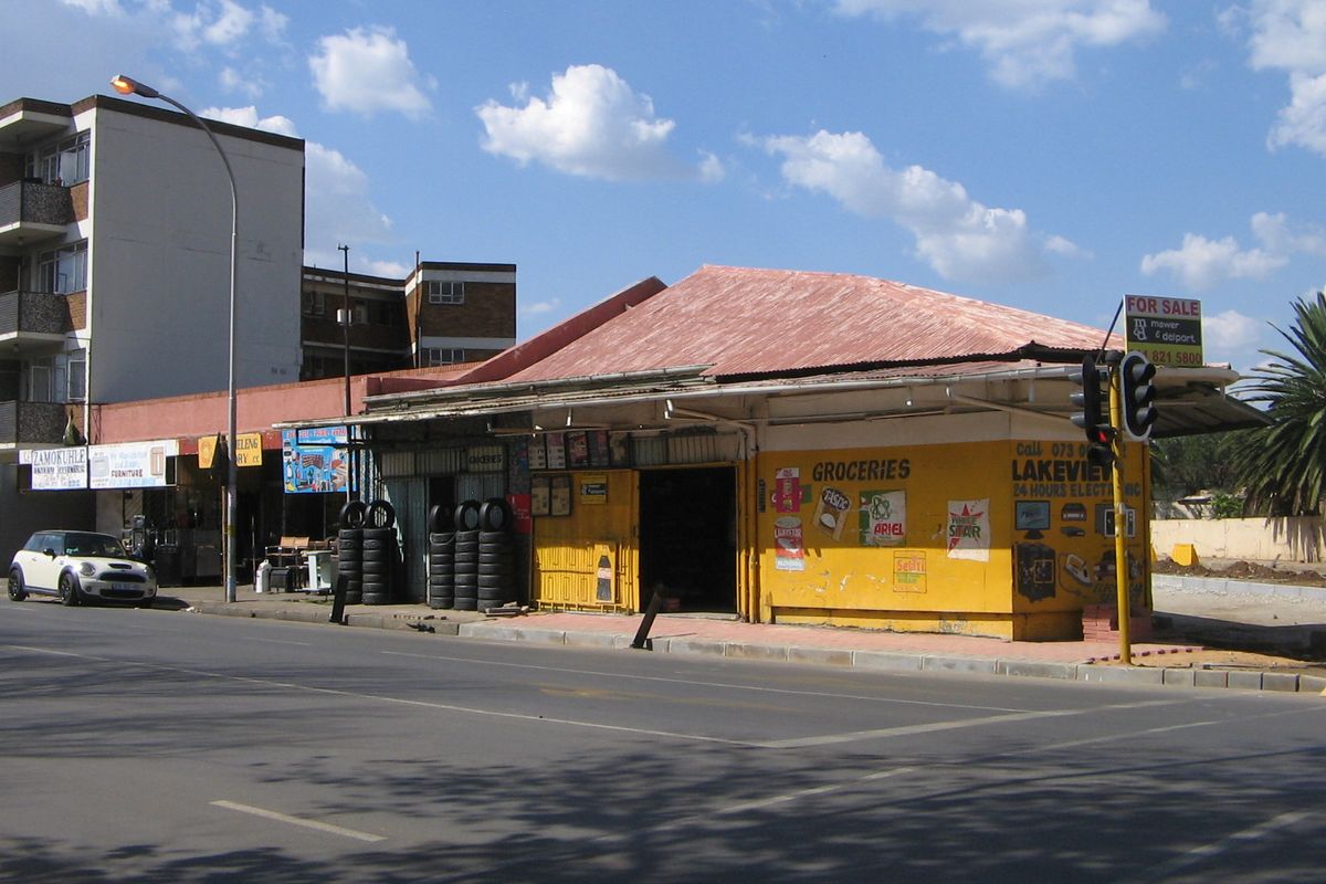 Street-level view of a retail property featuring a corrugated metal roof, yellow painted facade, and integrated security shutters along the sidewalk.