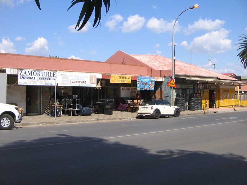 Street-level view of a retail strip featuring multiple shopfronts with metal security gates and signage along a public road.