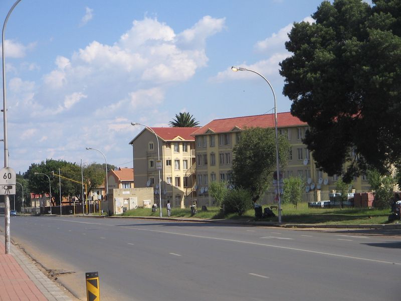 A street-level view of a multi-story residential apartment complex with tiled roofs, situated along a public road with sidewalk infrastructure.