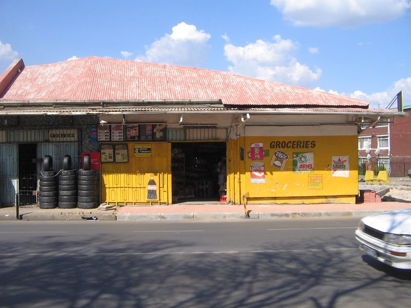 Street-facing retail storefront featuring a corrugated metal roof, yellow painted facade with signage, and secure metal roller shutter entrance.