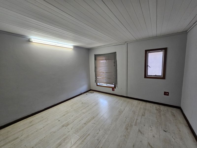 A vacant, neutral-toned office space featuring laminate flooring, a fluorescent light fixture, and a window with fitted venetian blinds.