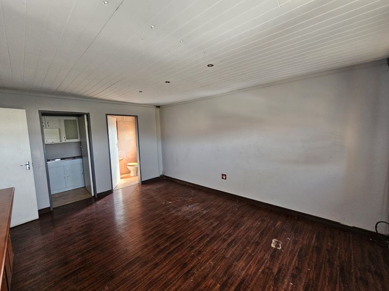 An empty living area featuring dark laminate flooring and a timber panel ceiling, with direct access to a small kitchenette and bathroom.