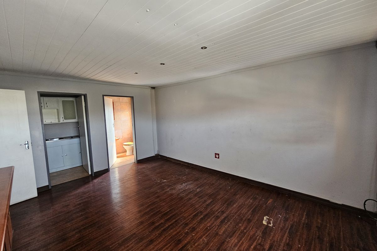 An empty living area featuring dark laminate flooring and a timber panel ceiling, with direct access to a small kitchenette and bathroom.