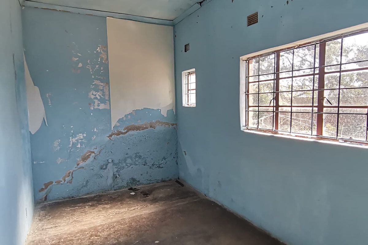 An empty bedroom requiring significant renovation, featuring damaged wall finishes, concrete flooring, and steel-framed windows with security bars.