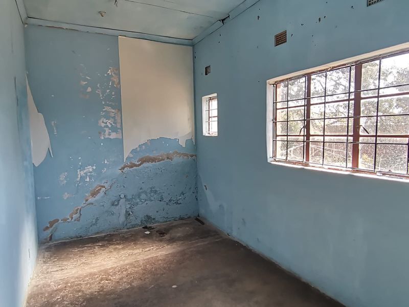 An empty bedroom requiring significant renovation, featuring damaged wall finishes, concrete flooring, and steel-framed windows with security bars.