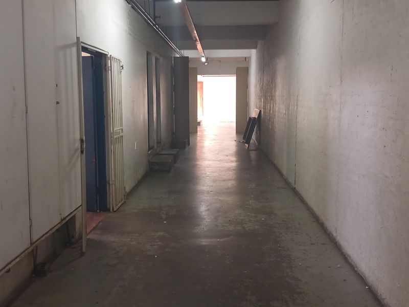 A long industrial hallway featuring concrete flooring, exposed conduit lighting, and security gate access, presenting a functional and utilitarian space.