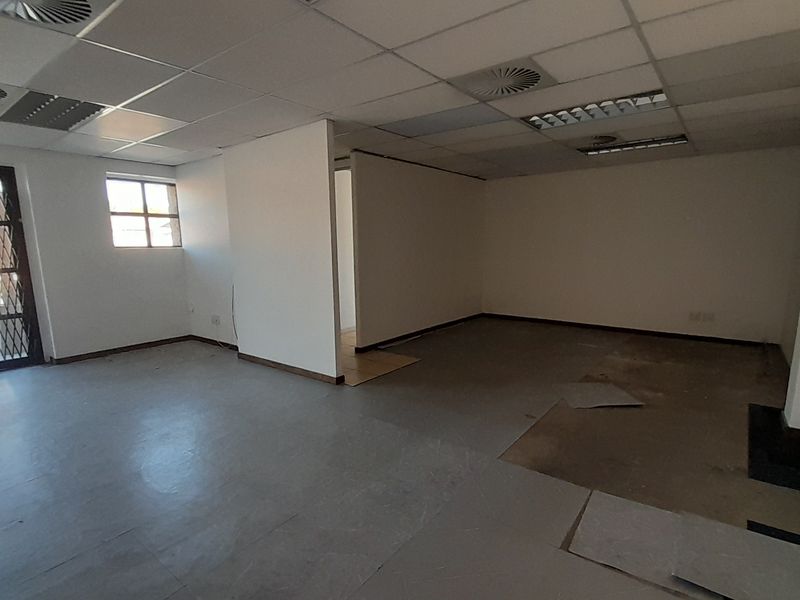 An empty office space featuring a suspended ceiling, recessed lighting, and a security gate, currently requiring flooring repairs.