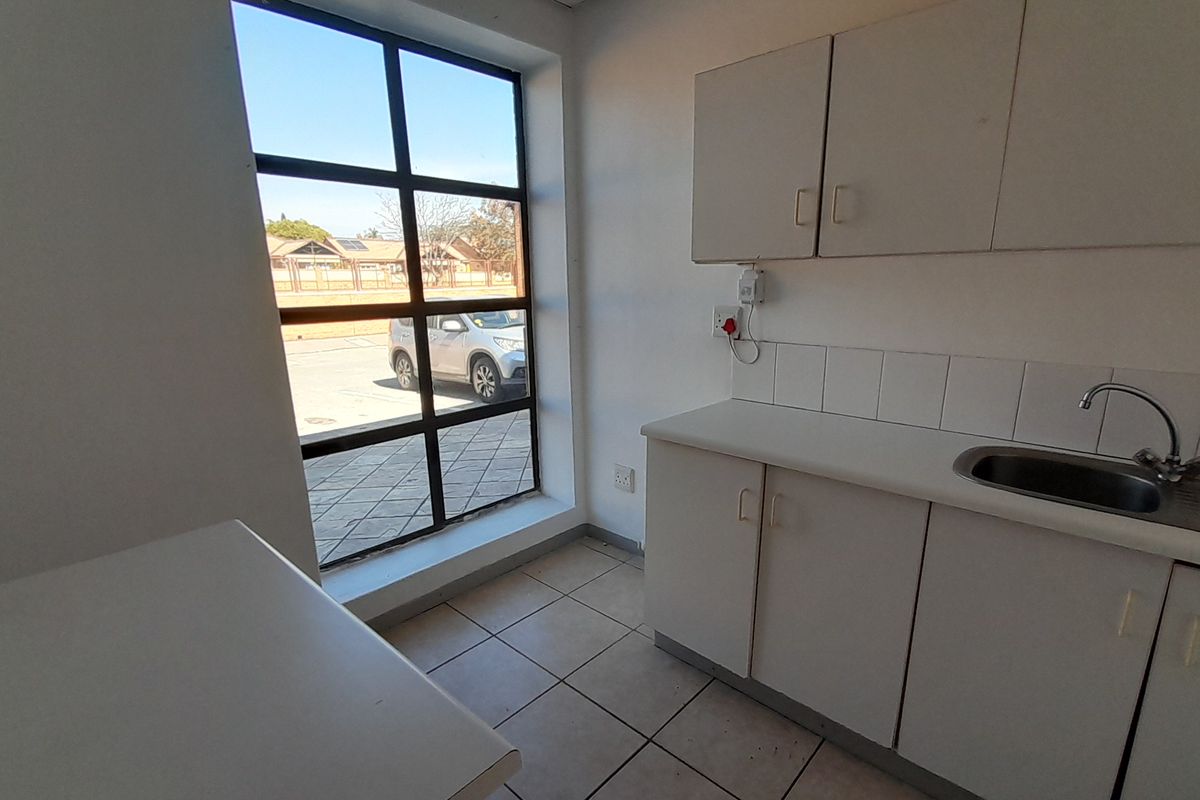 A compact, functional office kitchenette featuring built-in white cabinetry, a stainless steel sink, tiled backsplash, and large windows providing natural light.