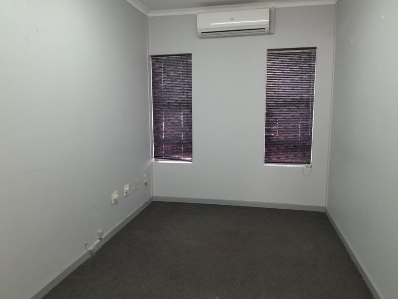 A clean, empty office space featuring grey carpeting, dual windows with venetian blinds, and a wall-mounted split-system air conditioning unit.