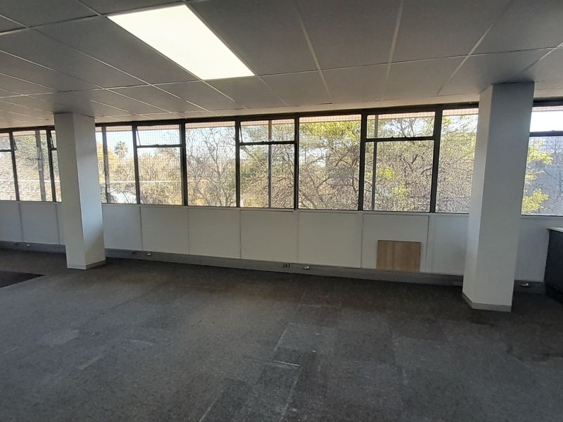 An open-plan office space featuring a suspended ceiling, large windows providing natural light, and a small built-in kitchenette area.