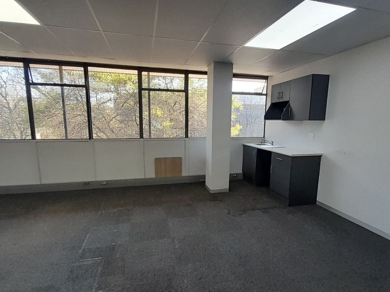 An empty office space featuring large windows, a small kitchenette area with built-in cabinetry, and a suspended ceiling with recessed lighting.
