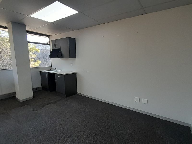An empty office space featuring a small kitchenette area, large windows providing natural light, drop ceilings, and carpeted flooring.