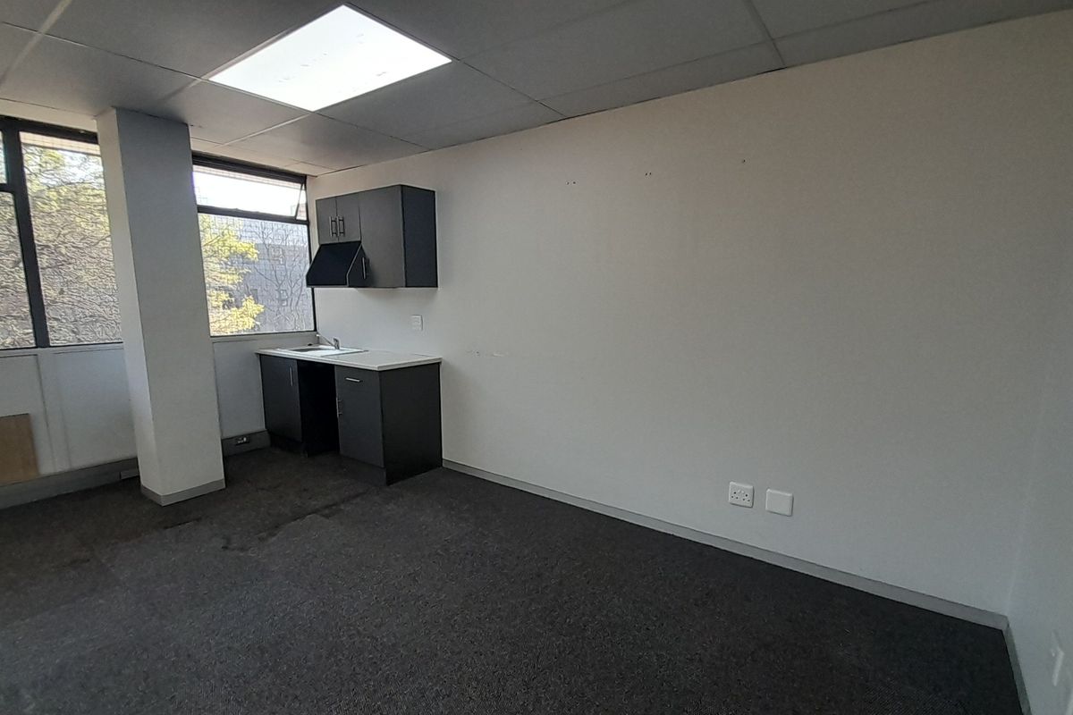 An empty office space featuring a small kitchenette area, large windows providing natural light, drop ceilings, and carpeted flooring.