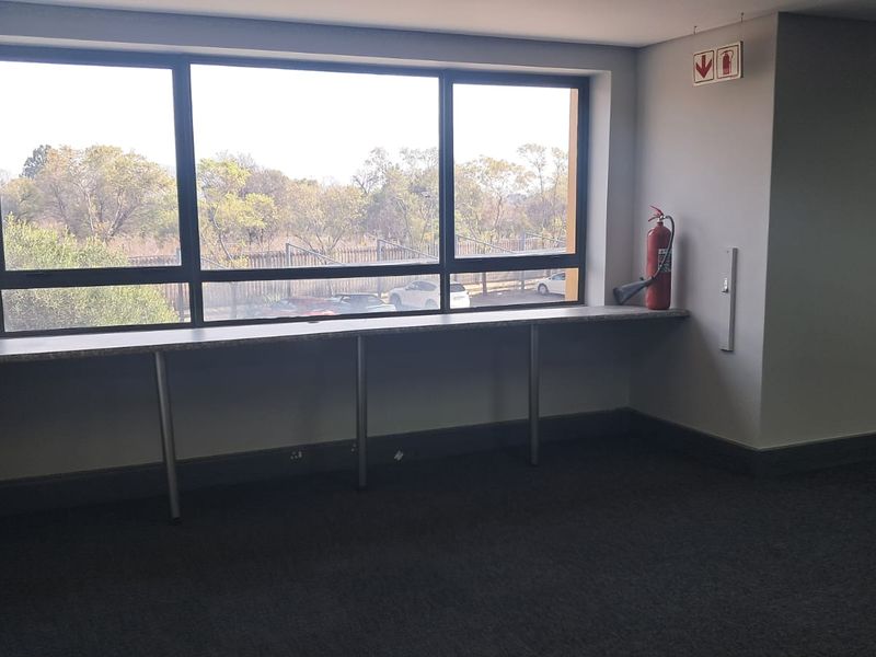 A bright, empty office space featuring a built-in desk along a large window, carpeted flooring, and essential fire safety equipment.