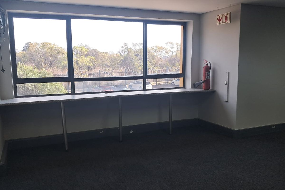 A bright, empty office space featuring a built-in desk along a large window, carpeted flooring, and essential fire safety equipment.