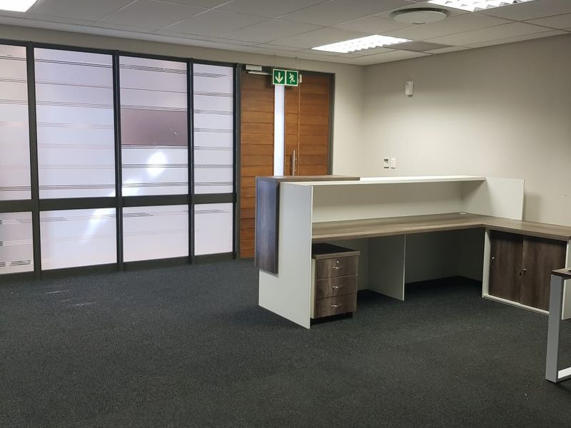 A professional office space featuring a built-in reception desk, frosted glass partitioning, suspended ceiling with recessed lighting, and dark carpet flooring.