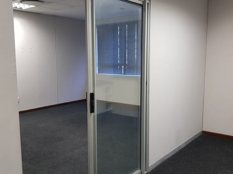 A clean, empty office space featuring a modern sliding glass door, suspended ceiling with recessed lighting, and neutral carpeted flooring.