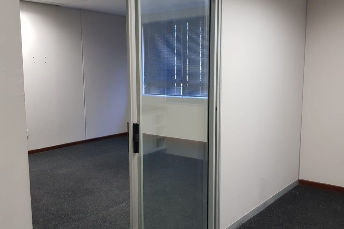 A clean, empty office space featuring a modern sliding glass door, suspended ceiling with recessed lighting, and neutral carpeted flooring.