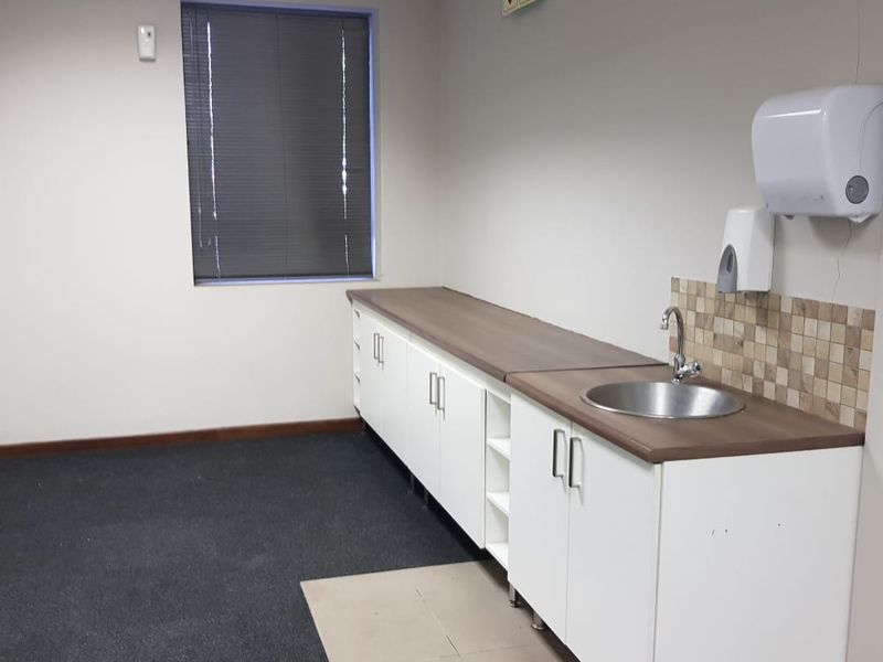 An office kitchenette area featuring built-in white cabinetry, a stainless steel sink, tiled backsplash, and carpeted flooring with a window and venetian blinds.