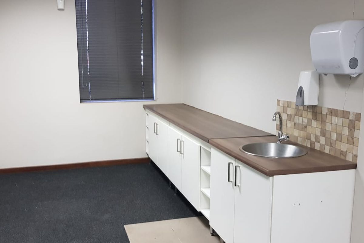 An office kitchenette area featuring built-in white cabinetry, a stainless steel sink, tiled backsplash, and carpeted flooring with a window and venetian blinds.