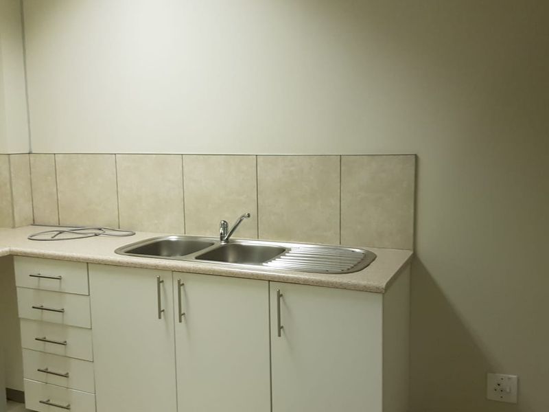 A clean, functional office kitchenette featuring built-in white cabinetry, a stainless steel double sink, and neutral tiled backsplash and flooring.