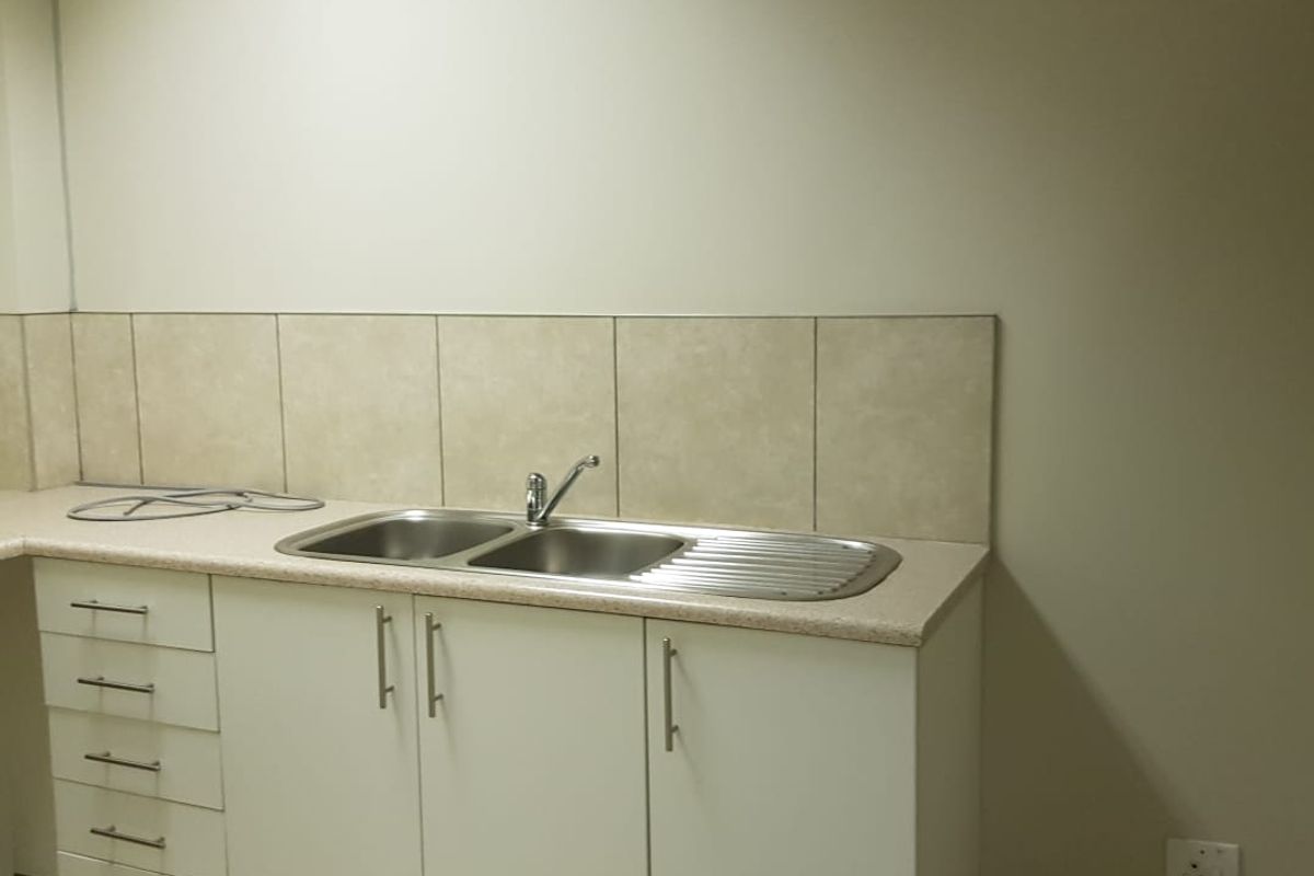 A clean, functional office kitchenette featuring built-in white cabinetry, a stainless steel double sink, and neutral tiled backsplash and flooring.