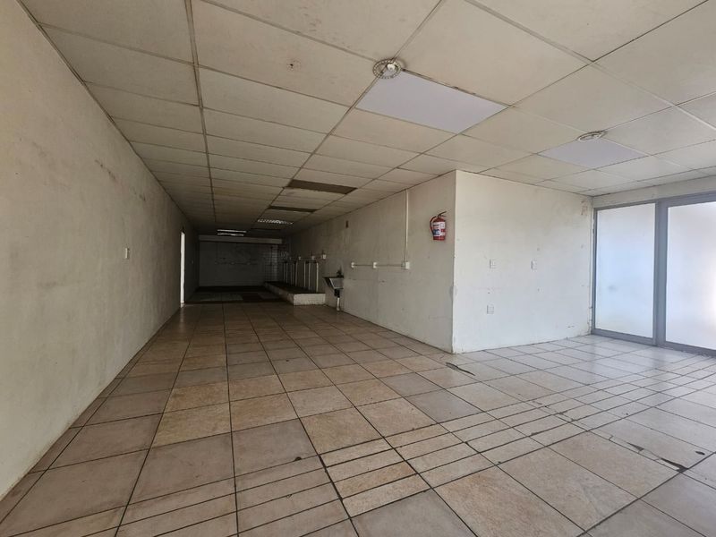 An empty, utilitarian retail space featuring ceramic tiled flooring, a suspended ceiling, and a wall-mounted sink, suitable for various commercial or storage purposes.