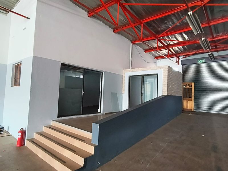 Spacious industrial utility area featuring exposed steel roof trusses, concrete flooring, a raised platform with sliding glass doors, and a secure roller shutter door.