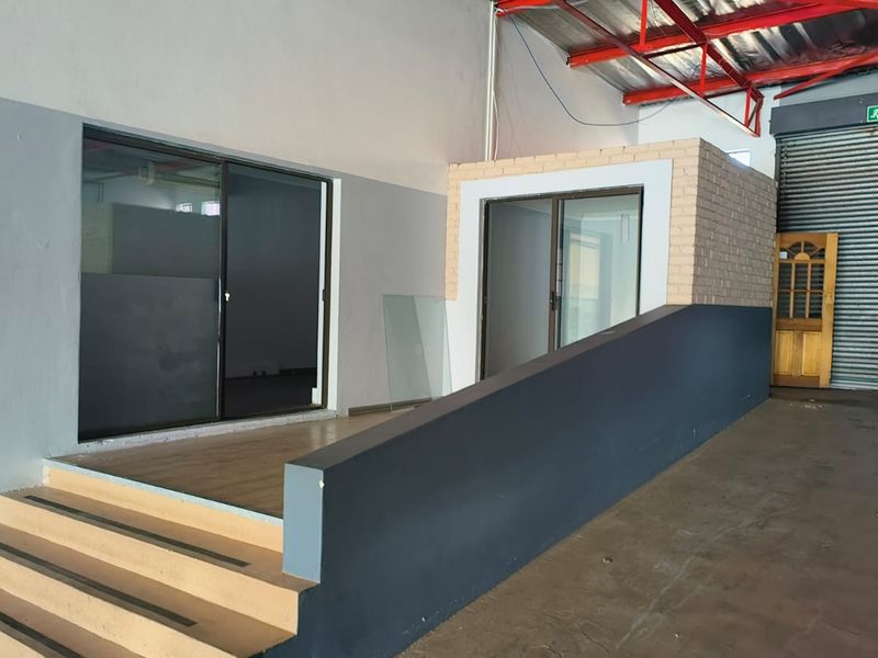 Industrial retail space featuring a roller shutter door, accessibility ramp, staircase, and partitioned office area with sliding glass doors.