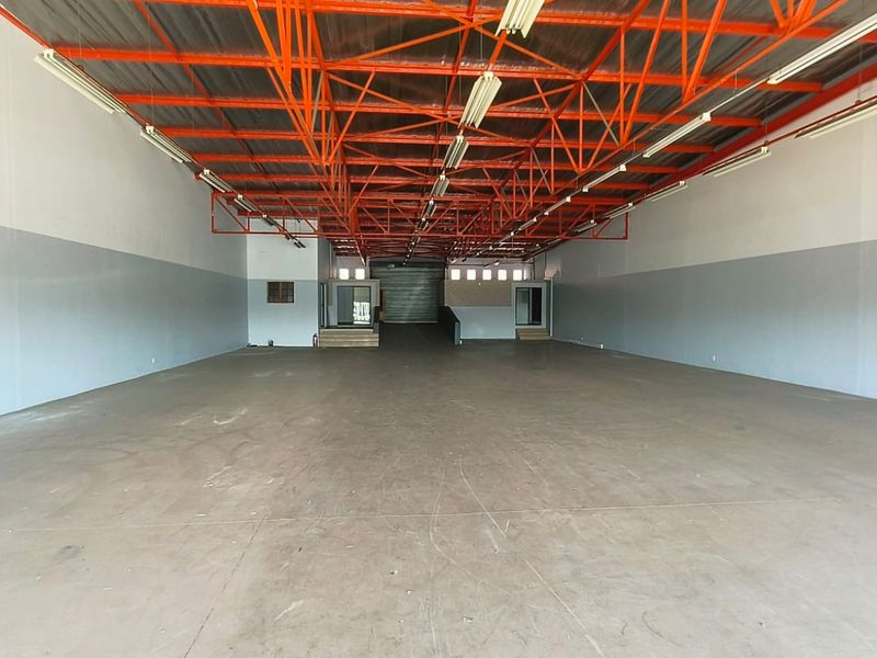 Spacious open-plan retail or warehouse area featuring high exposed steel roof trusses, concrete flooring, and a large roller shutter door for easy access.